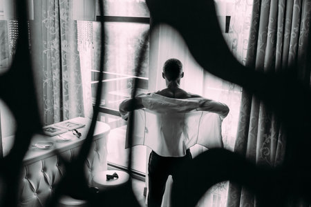 Muscular man gets ready for work by putting on his business shirt. Groom's morning preparation before wedding. Artwork. Back view. Black and white imageの写真素材
