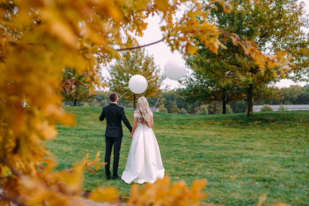 Happy bride and groom having a great time standing on the green grass with white balloons in their hands on their wedding day. Back viewの写真素材