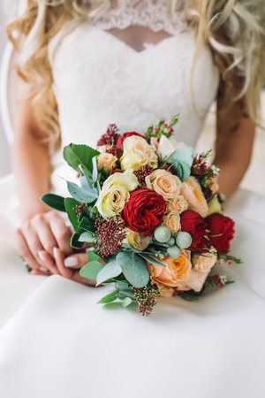 Bride holds a bunch of flowers. Beautiful bridal bouquet with white and peach roses, red peonies, and other flowers. Wedding morning. Close-up, indoor.の写真素材