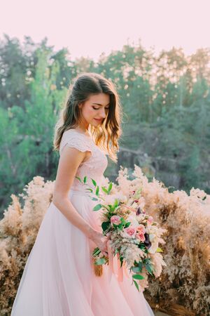 Bride with rustic wedding bouquet stands on the rock. Outdoor wedding ceremony, decorationの写真素材