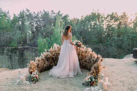 Bride with rustic wedding bouquet stands on the rock. Outdoor wedding ceremony. Back viewの写真素材
