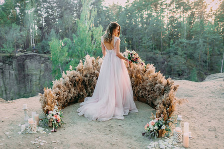 Bride with rustic wedding bouquet stands on the rock. Outdoor wedding ceremony. Back viewの写真素材