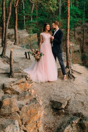 Groom is kissing his bride. Happy newlyweds embracing. Man in tuxedo and woman in a pink wedding dress is posing on nature. A ceremony outdoors. Close-up portraitの写真素材