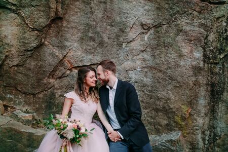 Attractive couple newlyweds bride and groom laughs and smiles to each other, happy and joyful moment. Man and woman in wedding clothes sit on the rock background. Outdoors.の写真素材
