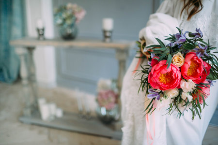 Bride holds in hands a rustic wedding bouquet with white roses and crimson peonies on window...