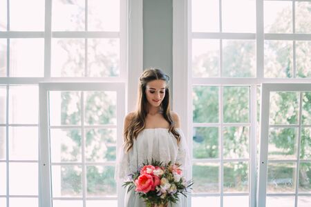 Bride holds in hands a rustic wedding bouquet with white roses and crimson peonies on window background. Close-up, indoorの写真素材