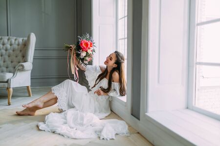 Cheerful, young bride holds a rustic wedding bouquet with peonies on panoramic window background. Close-up portrait. A happy brunette girl with a bunch of flowers.の写真素材