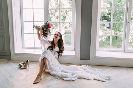 Cheerful, young bride holds a rustic wedding bouquet with white roses and crimson peonies on panoramic window background. Close-up portrait. A happy girl with a bunch of flowers., indoorの写真素材