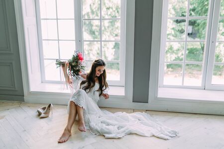 Cheerful, young bride holds a rustic wedding bouquet with peonies on panoramic window background. Close-up portrait. A happy brunette girl with a bunch of flowers.の写真素材
