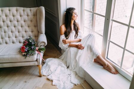 Woman by the window. Bride looking out the window, she waits for the groom. A bridal bouquet lies on the sofa. Indoorsの写真素材
