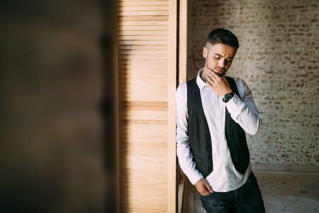 Thoughtful handsome guy in a white shirt and waistcoat near a window. Copy spaceの写真素材