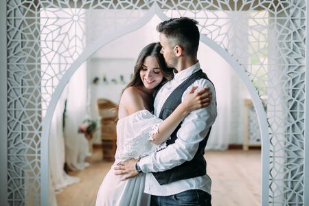 Cheerful young bride smiling and hugging groom while he kisses her in the head and embraces her waist. Beautiful newlyweds posing in vintage roomの写真素材