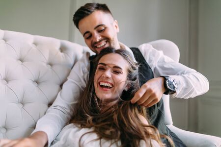A young couple in love having fun on a sofa and playing with a hair of the girl. Cheerful bride and groom laughing, close-up. Selective, soft focus on the girlの写真素材