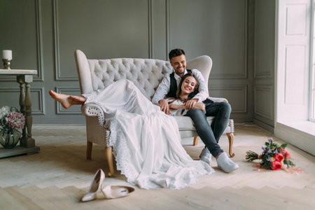 Young bride in tender lace dress lies on knees of a handsome groom in blue jeans. Cheerful newlyweds are laughing. Full length. Indoorsの写真素材