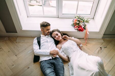 Cheerful bride and groom holding hands and lie on the wooden floor near the panoramic window. Wedding morning. Top viewの写真素材