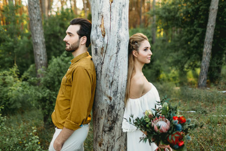 Bride and groom lean on the tree from different sides. Newlyweds are walking in the forest. Artworkの写真素材