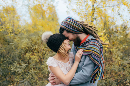 Bride in a knitted hat with a pom pom, hat covering her eyes. Groom in a colorful scarf. Happy young newlywed couple touching foreheads and noses in parkの写真素材