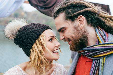 Bride in a knitted hat with pompom and groom with dreadlocks and scarf look at each other with tenderness and love. Close-upの写真素材