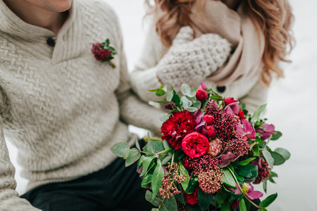 Bride and groom in warm winter clothes holds in hands a rustic wedding bouquet with red and crimson flowers. Close-up.の写真素材