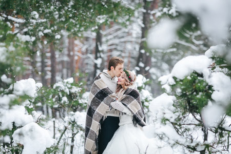 Couple in love tenderly hugs under the plaid on background of the snowy forest. Winter wedding.の写真素材