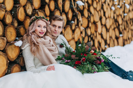Cheerful bride and groom with bouquet sits on snow on the wooden background. Winter wedding. Artworkの写真素材