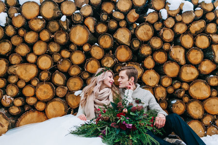 Bride and groom tenderly looks at each other. Stylish newlyweds sits on snow on the wooden background. Winter weddingの写真素材