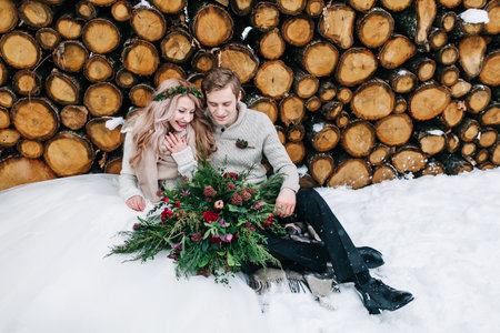 Bride and groom tenderly looks at each other. Stylish newlyweds with bouquet sits on snow on the wooden background. Winter weddingの写真素材