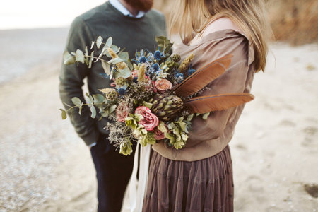 Newlyweds with rustic bouquet in hands stand on seashore. Bride and groom wear in country style clothes and hats. Outdoorsの写真素材
