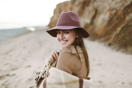 Portrait of cheerful lady in a hat with warm plaid stands on the beach. Woman walking along a seashore. Looking in camera Artwork. Close-upの写真素材
