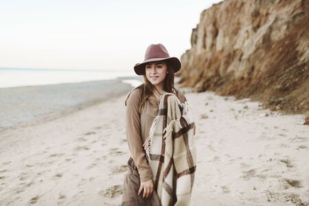 Satisfied girl in a hat with warm plaid admires the sea. Woman walking along a seashore. Artwork. Close-upの写真素材