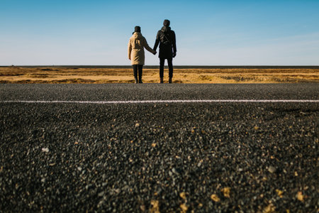 A young couple of travelers stands together on empty road on backgrouns of blue sky. Rear view....