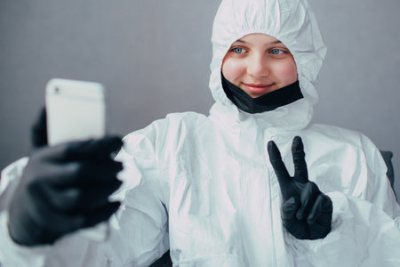 teenage girl in a viral protective suit with a black mask and  medical gloves is talking to friends on a video call because virologist is quarantined due the coronavirus epidemic.の写真素材