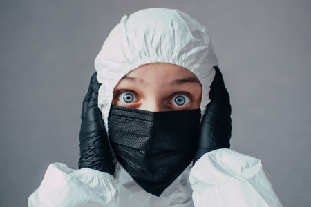 Virologist in black mask in a viral protective suit holds head, looks at the camera with fear and thinking about how to stop the coronavirus epidemic and what to do. Thinks about the dangers of this.の写真素材