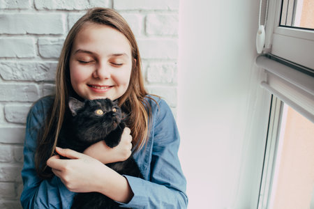 smiling girl with light brown hair give a hugs to black cat on a background of a white brick wall. Teenager closed eyes and cuddle a pet nearly window in new loft apartments.の写真素材