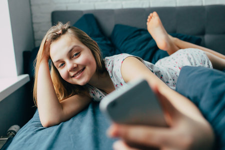 Young light brown hair woman in bed at home. Happy  girl smiling having  video call  on mobile telephone.  Teenager lying in bed and using cell phone for social media and internet.Lazy pajama morningの写真素材