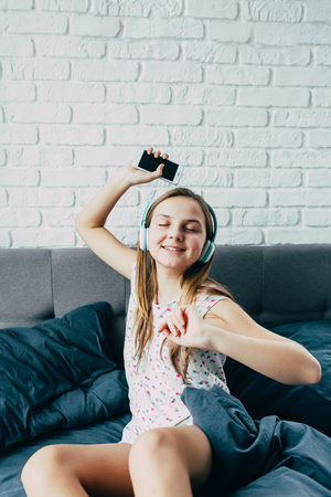 beautiful young Caucasian teenage girl sitting in bed with modern headphones is holding phone in her hand, dancing to cool music against large white wall. Active teenager morning. place for textの写真素材