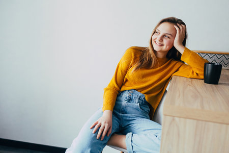 teenage girl in home interior looks left side, lean on wooden table with black cup of coffee and sitting on white modern chair. Beautiful girl in yellow sweater and blue jeans dreamingの写真素材
