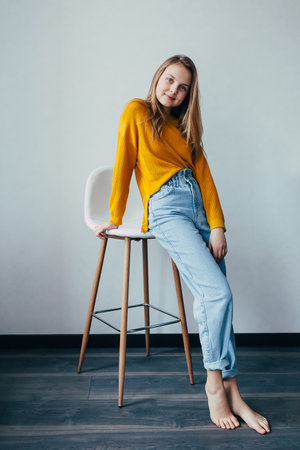 teenage girl looks at camera standing lean on white modern chair. Beautiful girl in yellow sweater and blue jeans in home interior with black wooden floor. Trendy casual outfit.の写真素材