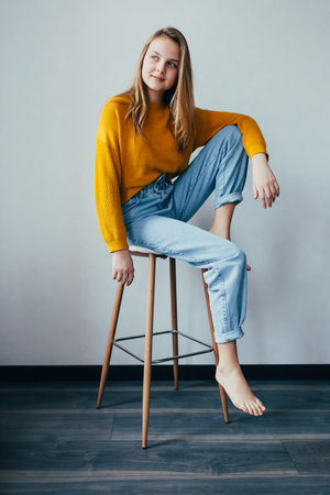 teenage girl looks at left sitting on white modern chair with one  leg raise up. Beautiful girl in yellow sweater and blue jeans in home interior with black wooden floor. Trendy casual outfit.の写真素材