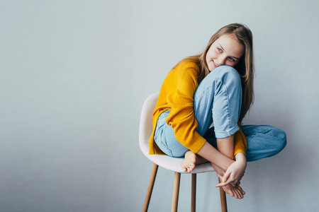 teenage girl looks at side and sitting on white modern chair with legs up. Beautiful girl in yellow sweater and blue jeans in home interior. Trendy casual outfit. place for textの写真素材