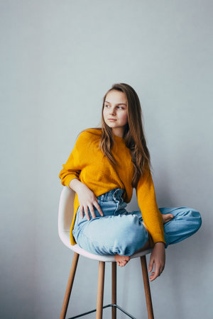 teenage girl looks at side sitting on white modern white chair with two leg raise up. Beautiful girl in yellow sweater and blue jeans in home interior. Trendy casual outfit.の写真素材