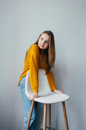 teenage girl looks at left side standing lean on white modern white chair. Beautiful girl in yellow sweater and blue jeans in home interior. Trendy casual outfit.の写真素材