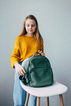 teenage girl looks on leather rucksacks and to fasten a backpack that lies on a white modern chair. Beautiful girl in yellow sweater and blue jeans stay in interior. Trendy casual outfitの写真素材