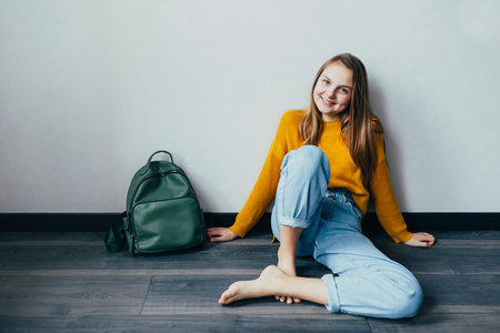 teenage girl looking on cellphone and smiles sitting on wooden floor. Beautiful girl in yellow sweater and blue jeans with backpack thrown nearby. Trendy casual outfit.の写真素材