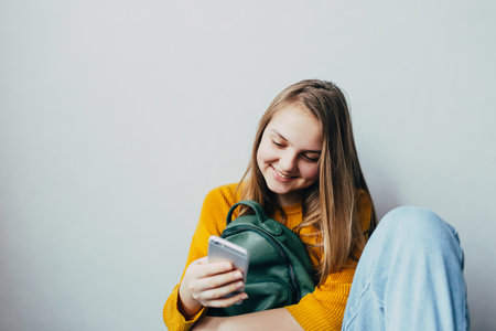 teenage girl looking on cellphone and smiles chatting with friends. Beautiful girl in yellow sweater and blue jeans holding mobile phone and cuddle green backpack. Trendy casual outfit. Place for textの写真素材