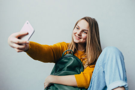 teen girl takes selfie on cellphone and smiles at camera. Beautiful girl in yellow sweater and blue jeans holding mobile phone and green backpack. Trendy casual outfit.の写真素材