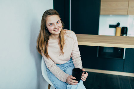 American teenage girl at Home kitchen interior holding Black coffee cup. Woman in pink blouse and blue jeans sitting on white chair. Modern interior designの写真素材