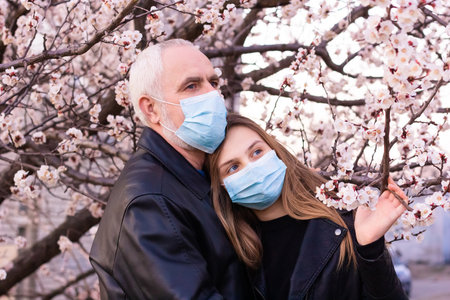 teenage girl and senior man  in face mask, standing in a flowering apricot blossom garden, support during pandemic and worry about  grandfather during world pandemic of coronavirus epidemic.の写真素材