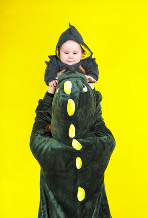 mom and daughter in a funny dragon costume. play on a yellow background. mother holds a little girl in a dragon costume over her head.の写真素材