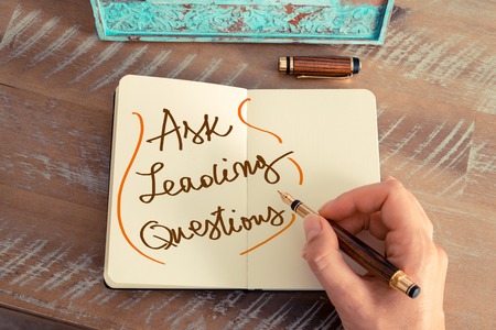 Retro effect and toned image of a woman hand writing a note with a fountain pen on a notebook. Handwritten text ASK LEADING QUESTIONS, business success concept Retro effect and toned image of a woman hand writing a note with a fountain pen on a notebook. Handwritten text ASK LEADING QUESTIONS, business success concept